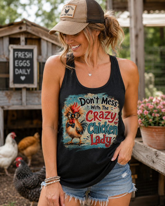 Woman wearing a black "Don't Mess With the Crazy Chicken Lady" racerback tank while standing in front of a backyard chicken coop with chickens. Funny chicken lover tank top by Woodland Willow Designs.