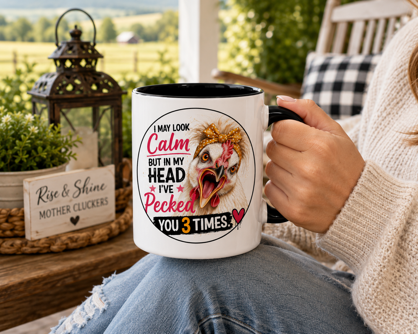 Person relaxing on a farmhouse porch holding a white 15 oz ceramic mug with black handle featuring Lucy the Silkie chicken and the saying "I May Look Calm But In My Head I've Pecked You 3 Times," with a cozy country landscape in the background.