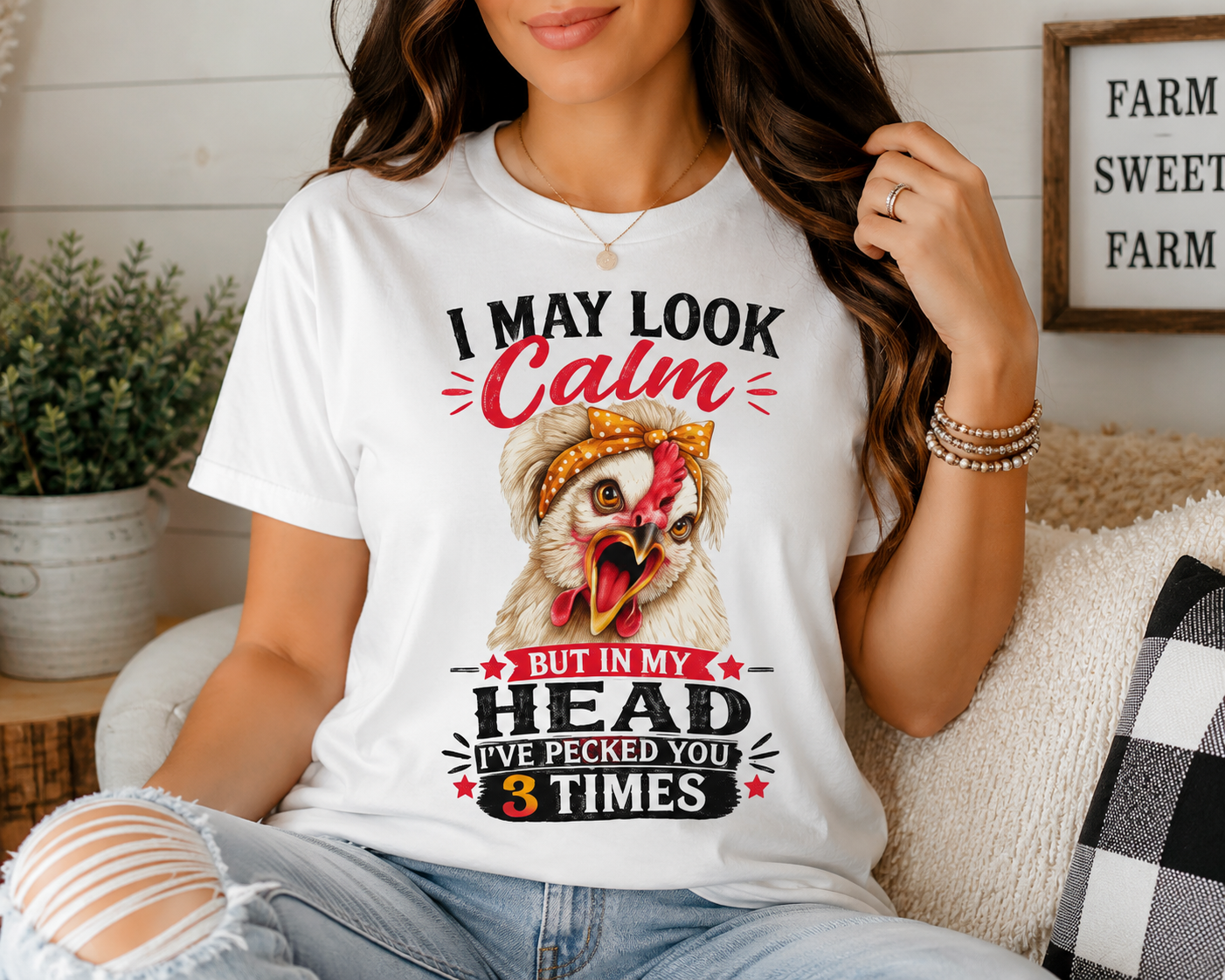 Woman wearing the "I May Look Calm" funny chicken graphic T-shirt by Woodland Willow Designs while relaxing on a cozy farmhouse sofa with a coffee mug. Features the Lucy & Friends™ design with the phrase "I May Look Calm... But In My Head I've Pecked You 3 Times," showcasing the shirt in a warm country-inspired lifestyle setting.
