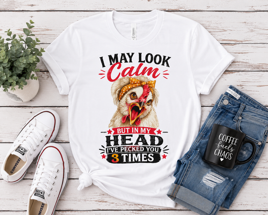 Flat lay of the "I May Look Calm" funny chicken graphic T-shirt by Woodland Willow Designs displayed with blue jeans, white sneakers, and farmhouse-inspired accessories on a rustic wood background. Features the Lucy & Friends™ design with the phrase "I May Look Calm... But In My Head I've Pecked You 3 Times."