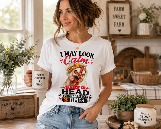 Woman wearing the "I May Look Calm" funny chicken graphic T-shirt by Woodland Willow Designs in a bright farmhouse kitchen while holding a coffee mug. Features the Lucy & Friends™ design with the phrase "I May Look Calm... But In My Head I've Pecked You 3 Times," showcasing the shirt in a cozy country lifestyle setting.