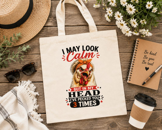 Flat lay of the "I May Look Calm" funny chicken canvas tote bag by Woodland Willow Designs displayed with a straw hat, sunglasses, notebook, coffee cup, and fresh flowers on a rustic wood background. Features the Lucy & Friends™ chicken design in a cozy farmhouse style.
